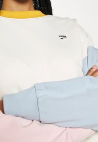 Sudadera de Puma con cuerpo crema, mangas azul claro y cuello amarillo. Logo destacado en el pecho y puños acanalados.