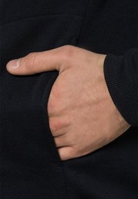 Hand of a person inserted into the front pocket of a black long-sleeve garment, showing wrist and part of the thumb.