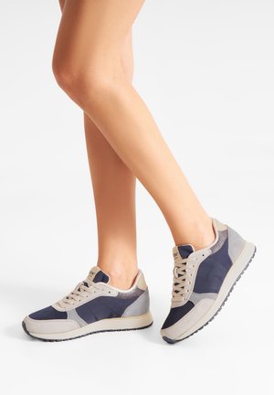 Ben iført beige og mørkeblå afslappede sneakers med snørebånd, stående med den ene fod fladt og den anden let løftet på en hvid baggrund.