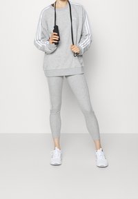 Grå idrottströja med vita ränder, grå tights och vita sneakers. Modellen håller i ett hopprep och visar en avslappnad passform och sportig design.