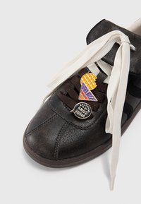 Baskets en cuir noir avec des lacets blancs et bruns, ornées de pin's en forme de cornet de glace et d'une bulle de dialogue disant "BIENVENUE À L'ENVERS".