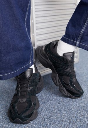 Black sneakers featuring a combination of textured and smooth materials, prominent lacing, and a chunky sole, paired with dark denim pants.