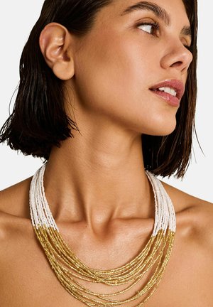 Femme aux cheveux courts et foncés portant un collier multi-rangs de perles blanches et dorées, regardant sur le côté contre un fond uni.