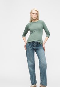 Ξανθιά γυναίκα στέκεται με τα χέρια στις τσέπες, φορώντας πράσινη μπλούζα με μακρύ μανίκι, ψηλόμεσο μπλε τζιν με δέσιμο μπροστά και παπούτσια με σχέδιο.