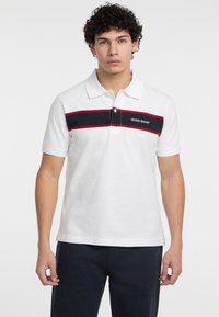 Camisa polo blanca de manga corta, con una franja horizontal negra y roja en el pecho y el logo de "Guess Jeans" en negro.