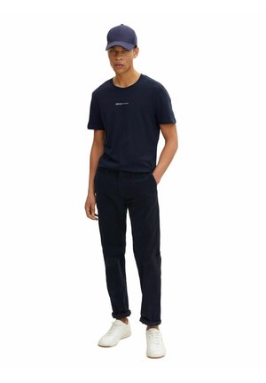 Marineblaue Baumwoll-T-Shirt mit dem Schriftzug "DENIM", dunkle cordfarbene Hose und weiße Sneaker. Die Mütze ist ebenfalls marineblau mit einem gebogenen Schirm.