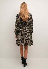 Schwarz-beige gemustertes Kleid mit langen Ärmeln, tailliertem Bund und ausgestellt Rock. Trägt schwarze Knöchelstiefel.