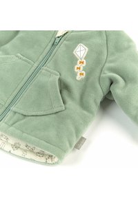 Giacca in pile verde con zip frontale, dettaglio ricamato a forma di aquilone e fodera decorata con piccole illustrazioni di animali.