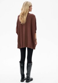 Brązowy, oversize sweter z dzianiny, z opadającymi ramionami, prążkowanymi mankietami i dołem, w połączeniu z czarnymi legginsami i błyszczącymi czarnymi muszkieterkami do kolan.