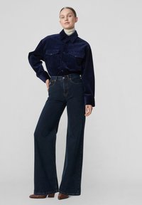 Nő sötétkék kord ingben, fehér garbóban, magasderekú, széles szárú sötét farmernadrágban és barna hegyes orrú csizmában áll szürke háttér előtt.