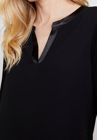 Blouse noire avec un décolleté en V et un bord en satin au col. Elle présente une texture douce et lisse ainsi que des manches longues.