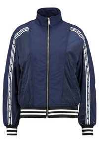 Námořnická modrá bomber bunda s vysokým límcem, zapínáním na zip, elastickými manžetami a lemem. Má bílé boční pruhy s názvem značky. Hladká textura.