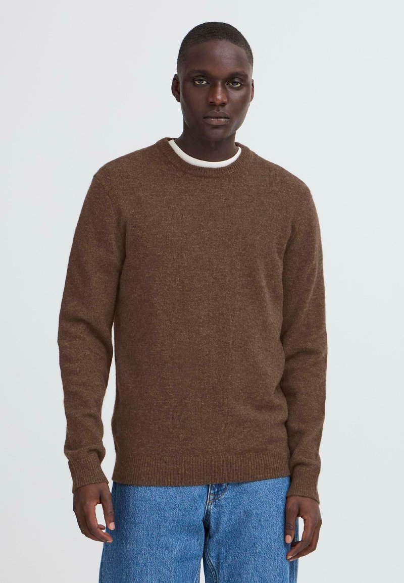 Brauner Strickpullover mit rundem Halsausschnitt, gerippten Bündchen und Saum; über einem weißen Hemd mit Kragen getragen; kombiniert mit hellblauen Jeans.