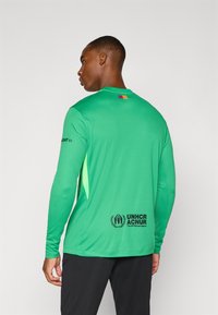Maglietta verde a maniche lunghe con scollo rotondo, dotata di una piccola bandiera sulla parte posteriore e un logo sul lato inferiore, realizzata in tessuto testurizzato.