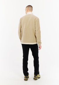 Man met zijn rug naar de camera, gekleed in een beige fleece jacket, zwarte broek en gele wandelschoenen, staand op een witte achtergrond.