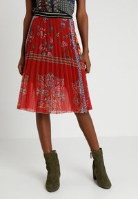 Rode geplooide rok met bloem- en geometrische patronen, voorzien van een aangesloten tailleband en decoratieve accenten. Gecombineerd met groene enkellaarsjes.