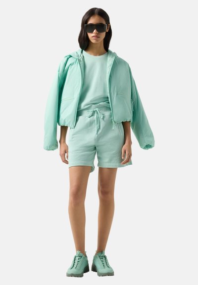 Femme portant une doudoune oversize vert menthe, sweat-shirt assorti, short, baskets épaisses et grandes lunettes de soleil noires, debout devant un fond blanc.