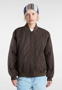Braune gesteppte Bomberjacke mit geripptem Kragen und Bündchen, die eine glatte Textur und subtile wellenförmige Muster auf dem Stoff aufweist.