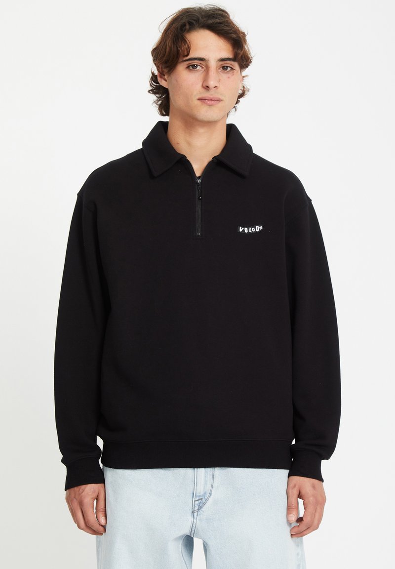 Pull en polaire noir avec un col zippé et un logo sur la poitrine gauche ; doté de poignets et d'une ceinture côtelés, associé à un jeans bleu clair.
