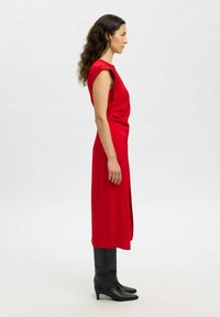 Frau mit langen,lockigen Haaren, die ein ärmelloses rotes Midikleid und schwarze kniehohe Stiefel mit mittelhohem Absatz trägt, im Profil vor einem weißen Hintergrund stehend.