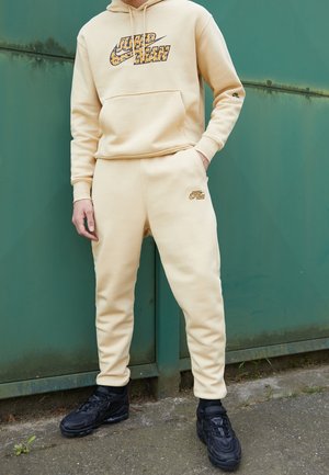 Person trägt beige Nike Hoodie und Jogginghose mit Leopardenmuster-Logo, steht vor einer grünen Metalltür, schwarze Sneakers auf dem Bürgersteig.