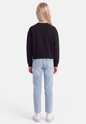 Enfant aux longs cheveux blonds portant un sweat-shirt noir, un jean bleu clair et des baskets blanches montantes, debout de dos sur un fond blanc.