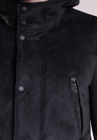 Czarna zamszowa kurtka z wysokim kołnierzem, posiadająca cztery guziki oraz kieszeń zapinaną na zamek po lewej stronie. Miękka w dotyku i elegancka w designie.