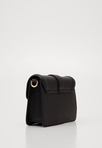 Svart strukturerad lädercrossbodyväska med lockstängning och ring i guldfärgad metall på en neutral bakgrund.