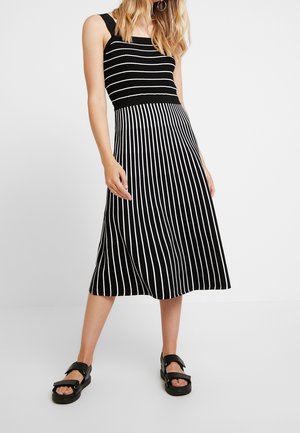 Donna che indossa un vestito nero senza maniche con righe bianche orizzontali nella parte superiore e righe verticali nella gonna, abbinato a sandali neri.