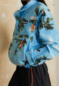 Person wearing a blue Columbia jacket with floral and bird prints, and black pants, standing against a textured beige wall.