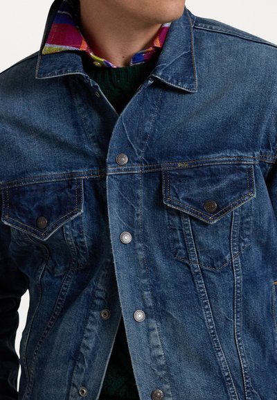 Denimjakke med lange ermer, to brystlommer, knappestup foran og klassisk krage. Har en mørkeblå farge og merkbar sømdetalj.