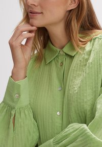 Hellgrüne, strukturierte Bluse mit Kragen, Knöpfen und puffärmel. Sie hat vertikale Streifen und Ausstanzungen an den Ärmeln.