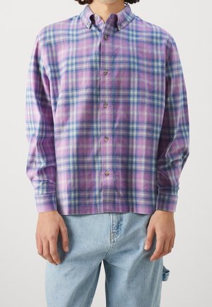 Hombre con camisa de manga larga abotonada con patrón a cuadros rosa, azul y blanco, y pantalones vaqueros azul claro.