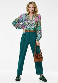 Bunt gemusterte Bluse mit Ballonärmeln, grüne hochtaillierte Hose, brauner Gürtel und braune Handtasche mit gedrehtem Tragegurt, schwarze Schuhe.