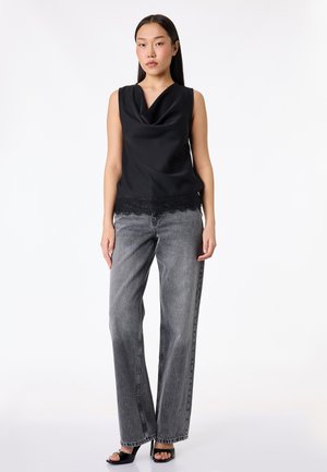 Kvinde iført sort satin top uden ærmer med blondekant, grå vidde-benet jeans og sorte åbne tå-sandaler, stående mod hvid baggrund.