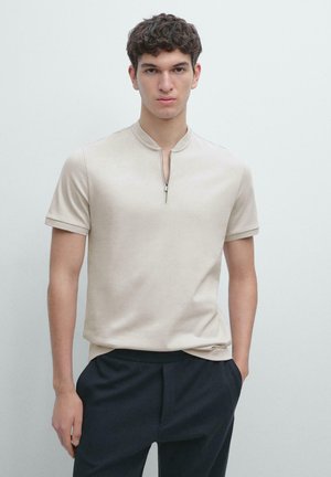 Młody mężczyzna w beżowej koszuli z krótkim rękawem zapinanej na zamek, włożonej do czarnych spodni, stojący na tle jasnego, jednolitego tła.