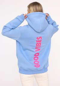 Hoodie bleu clair avec un grand graphisme "GOOD VIBES" en rose au dos. Présente une capuche avec cordon de serrage et des poignets et un ourlet côtelés.