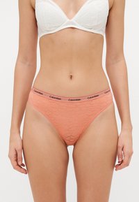 Őszibarack színű csipkés tangás alsó fekete Calvin Klein derékkal. Virágmintával és sima textúrával rendelkezik, amely a testhez simuló illeszkedésre lett tervezve.