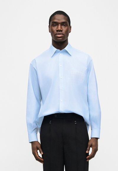 Άνδρας που φοράει ανοιχτό μπλε πουκάμισο με μακριά μανίκια και μαύρο παντελόνι, στέκεται μπροστά σε απλό λευκό φόντο.