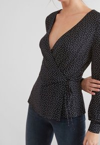 Blouse noire à pois blancs, avec un décolleté en V profond, des manches longues et un lien à la taille. Texture de tissu lisse.