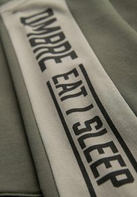 Sudadera verde con una banda de crema bordada que lleva el texto negro "OMBREEAT/SLEEP" en diferentes estilos de fuente. Textura suave con un diseño casual.