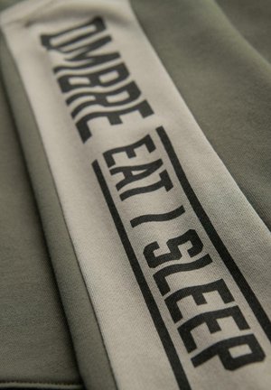 Groene sweatshirt met een gestikte crèmekleurige band waarop in zwarte tekst "OMBREEAT/SLEEP" in verschillende lettertype-stijlen is geschreven. Zachte textuur met een casual ontwerp.