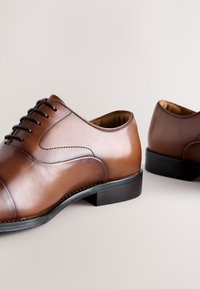 Paire de chaussures habillées en cuir marron poli avec lacets noirs et talons bas empilés sur un fond neutre.