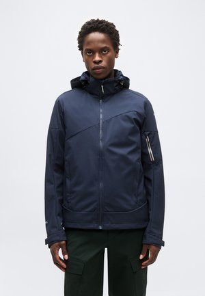 Άνδρας φοράει μπορντό σακάκι με κουκούλα, φερμουάρ μπροστά και στα μανίκια, στέκεται μπροστά σε ένα μονόχρωμο γκρι φόντο.