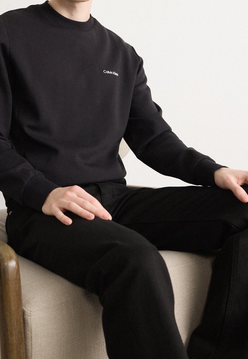 Felpa nera con colletto tondo e logo "Calvin Klein" bianco, in materiale misto cotone, con polsini a coste, indossata con pantaloni neri, seduto su una sedia chiara.