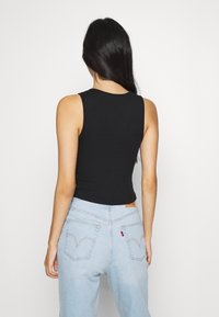 Kobieta z długimi czarnymi włosami, ubrana w bezrękawnik w kolorze czarnym i jasnoniebieskie spodnie o wysokim stanem, stoi przodem do białego tła.