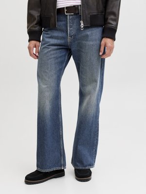 Man draagt vervaagde blauwe wijduitlopende spijkerbroek, zwarte riem, zwarte leren jas en zwarte schoenen, staand tegen een effen achtergrond.