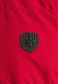 Roter Stoff mit einem strukturierten schwarzen Schild-Emblem, das einen zentralen Stern und dekorative Nieten zeigt, vor einem glatten Hintergrund platziert.