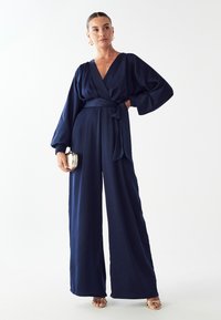 Navyblauwe jumpsuit met een diepe V-hals, lange ballonsleeves en een getailleerde taille. Zachte, gladde stof met een wijd uitlopende pijp.