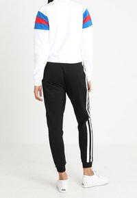Vit zip-up jacka med röda och blå ränder på ärmarna, matchad med svarta joggers med vita sidränder och vita sneakers.